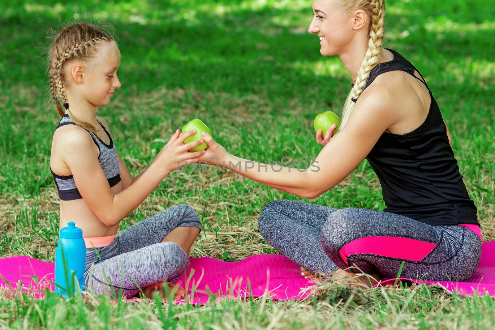 Mother is giving to daughter a green apple sitting on the roll mat in park.