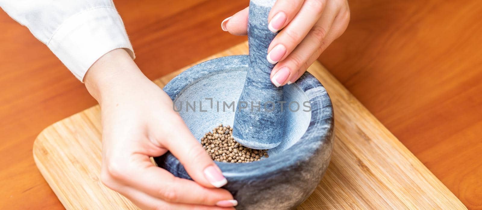 Hands of woman grinding black pepper by pestle in a gray granite mortar on the table.