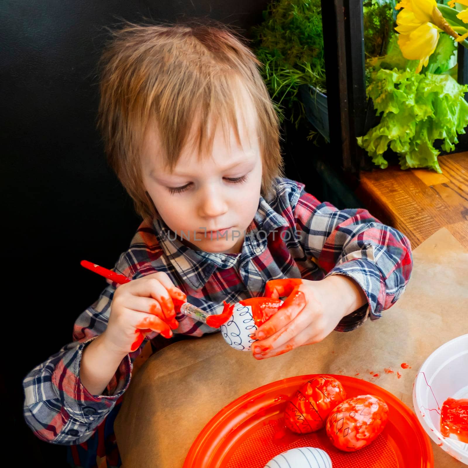 funny kid in a red plaid blouse paints artificial Easter eggs red on a wooden table among fresh greenery. preparation for the bright Easter holiday