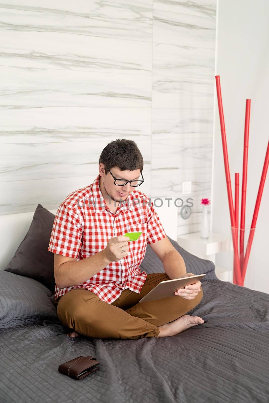 Online shopping concept. Young man sitting on the bed and shopping online using tablet, looking at the credit card