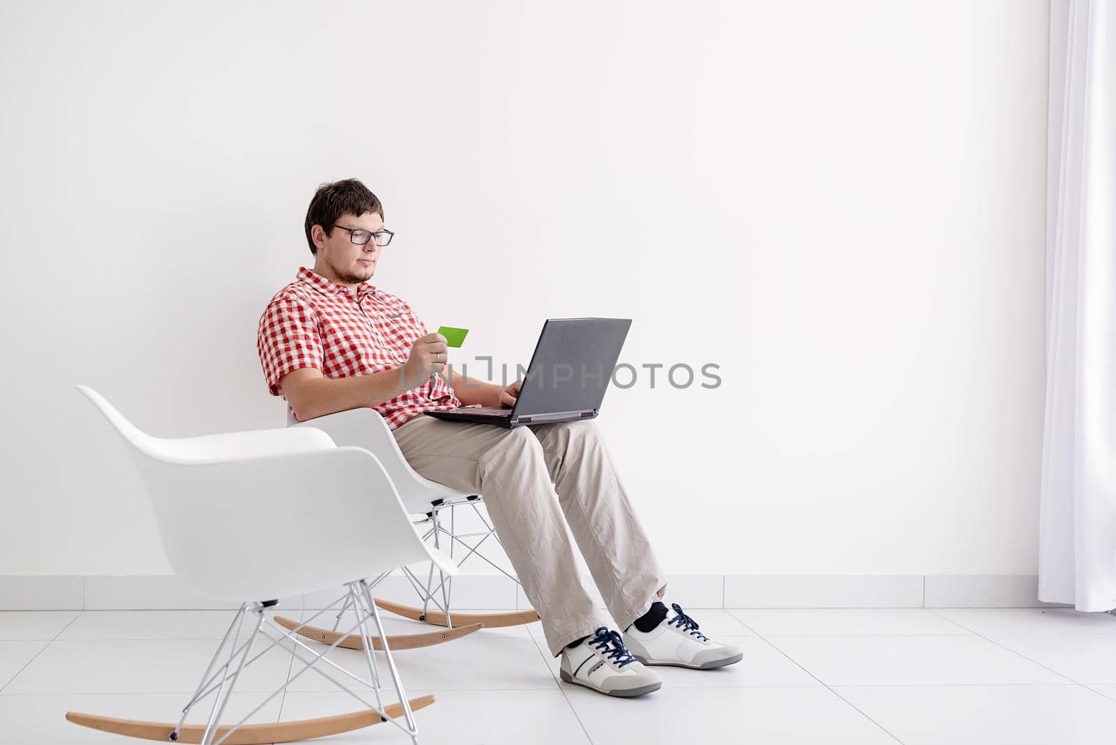 Online shopping concept. Young man sitting in the chair and shopping online using laptop looking at the credit card, copy space