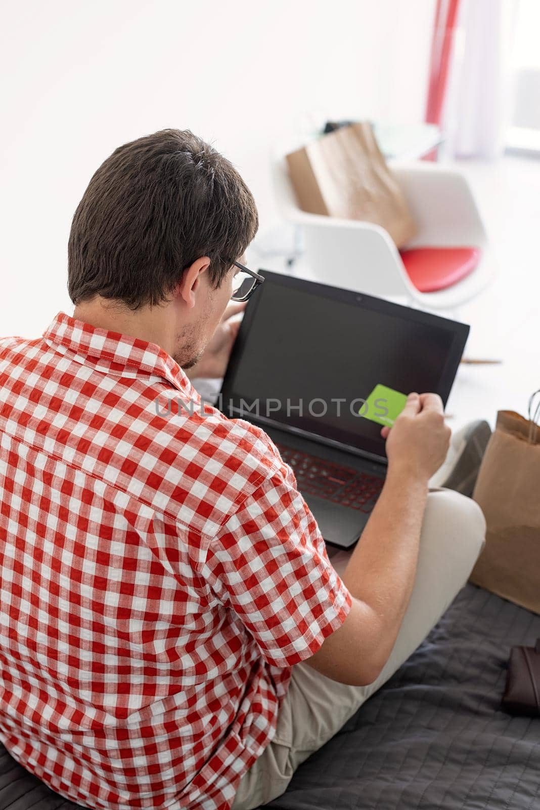 Online shopping concept. Young man sitting on the bed and shopping online using laptop looking at the creadit card, blank screen