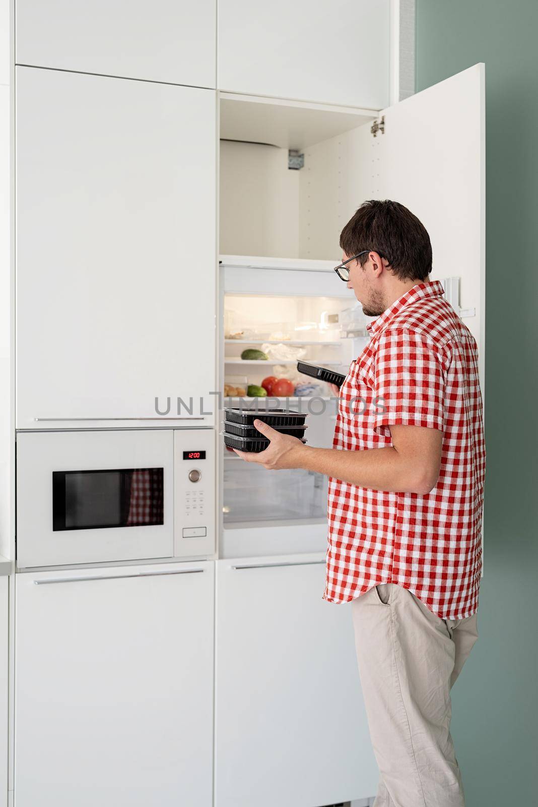 Delivery food, products to home. Shopping and healthy food concept. Young man in red plaid shirt holding disposable plastic boxed with food and putting them to the refridgerator