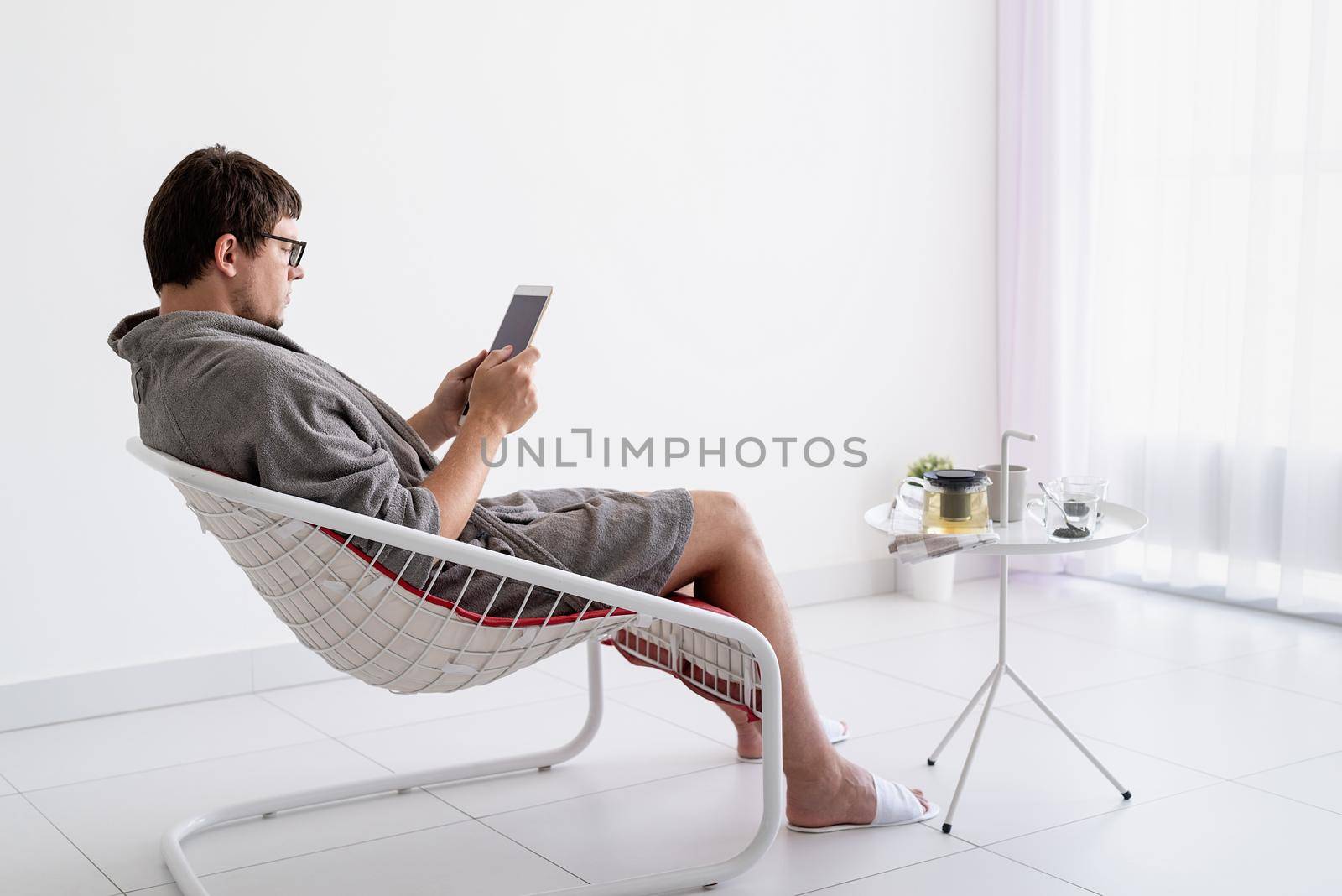 man siting in bathrobe drinking tea and using tablet at home by Desperada