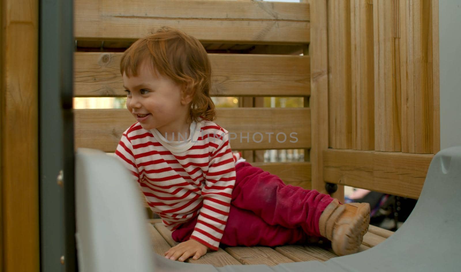 Little girl sitting on a top of a wooden slide and smiling. Baby on the playground in the park.