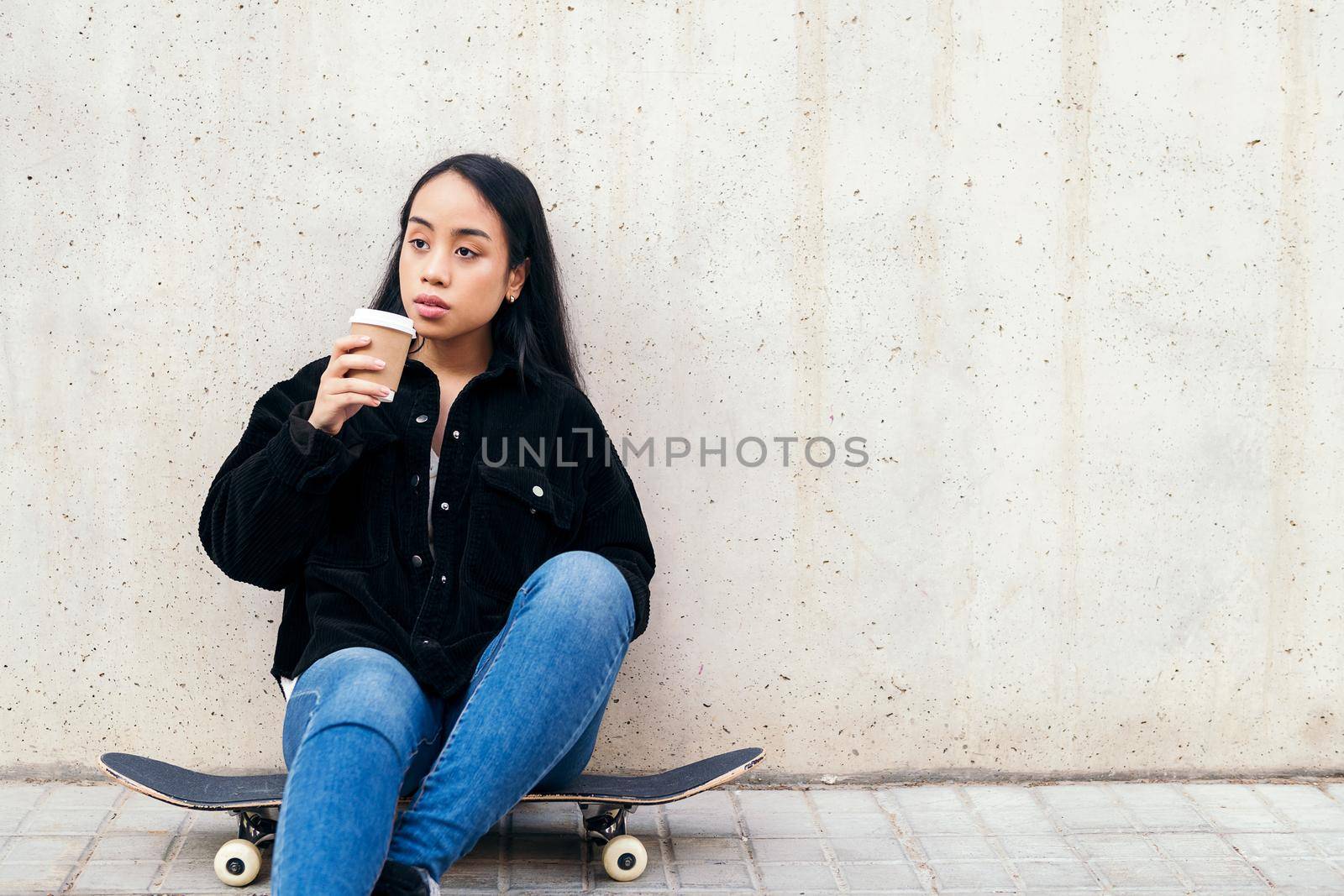 young asian woman drinking coffee outdoors sitting on her skateboard leaning against a concrete wall