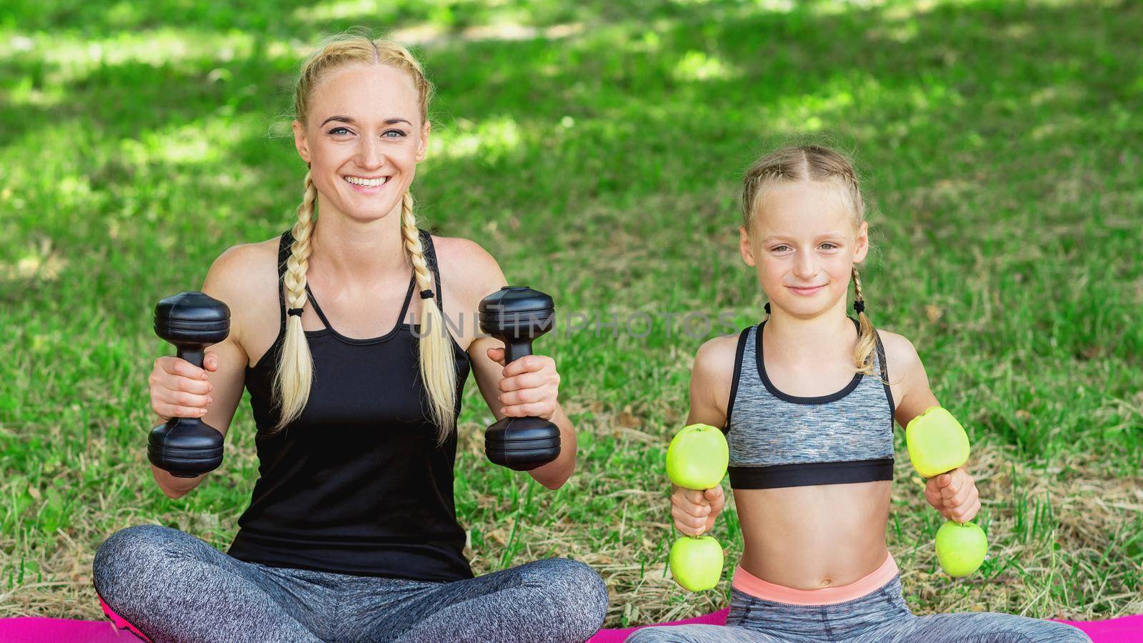 Young mother with her little daughter are training with apple dumbbells in the park.