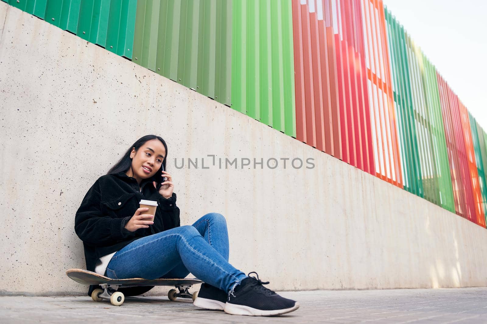 asian teenage girl talking by phone outdoors sitting on her skateboard leaning against a concrete wall