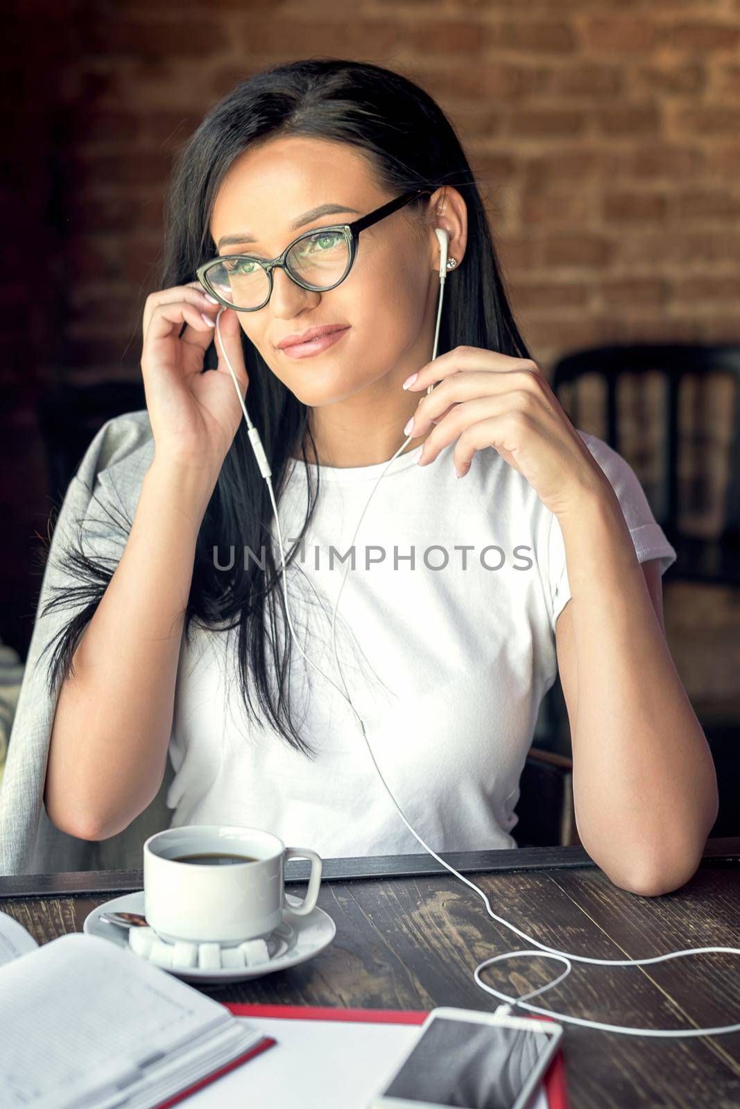 Beautiful smiling woman in glasses is listening music by her smartphone at cafe. by okskukuruza