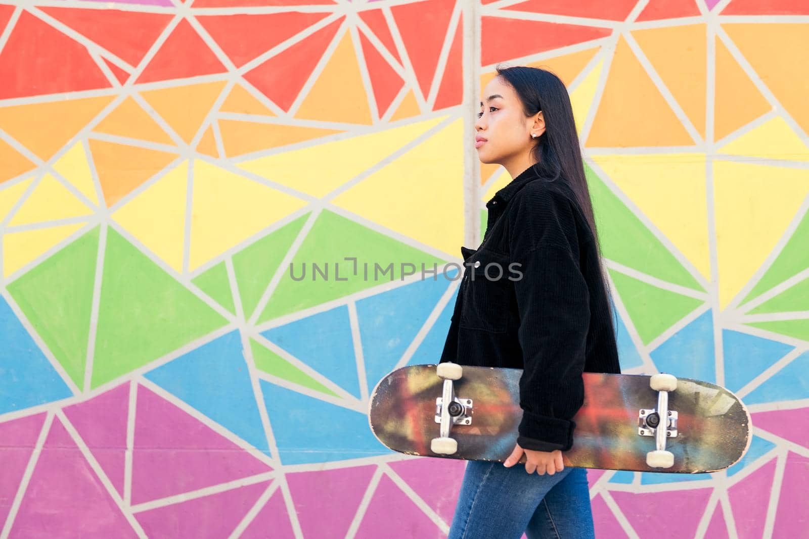 young asian woman walks with a skateboard in her hand in front of a colorful wall