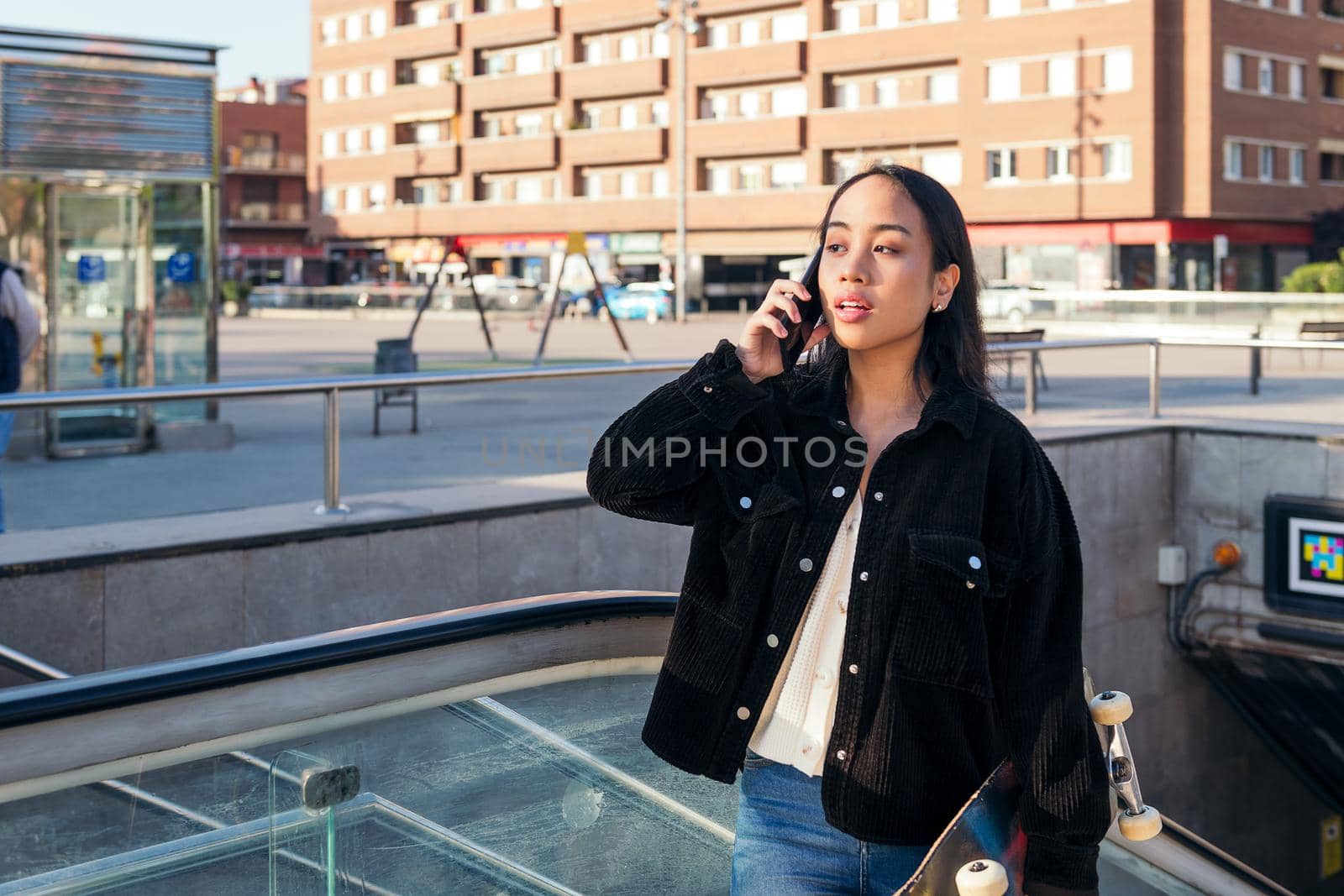 young asian woman exits subway station with skateboard in hand while talking on phone