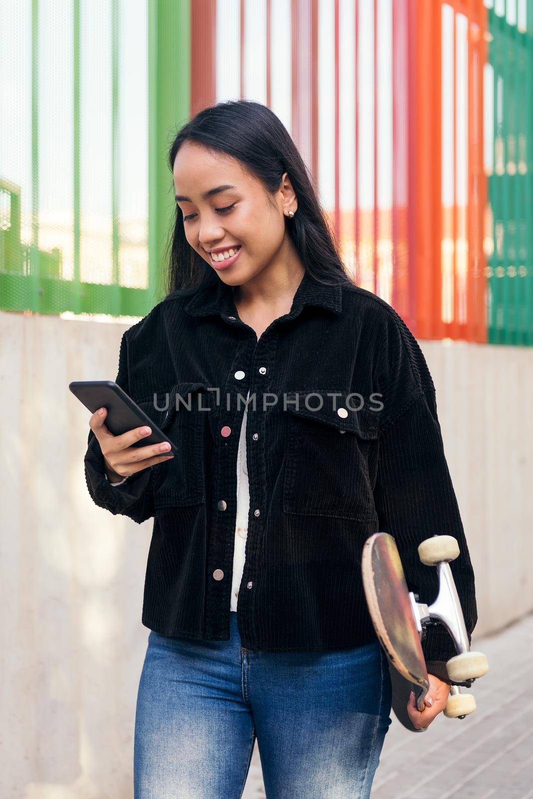 happy asian girl typing on the mobile phone while walking down the street with her skateboard in her hand