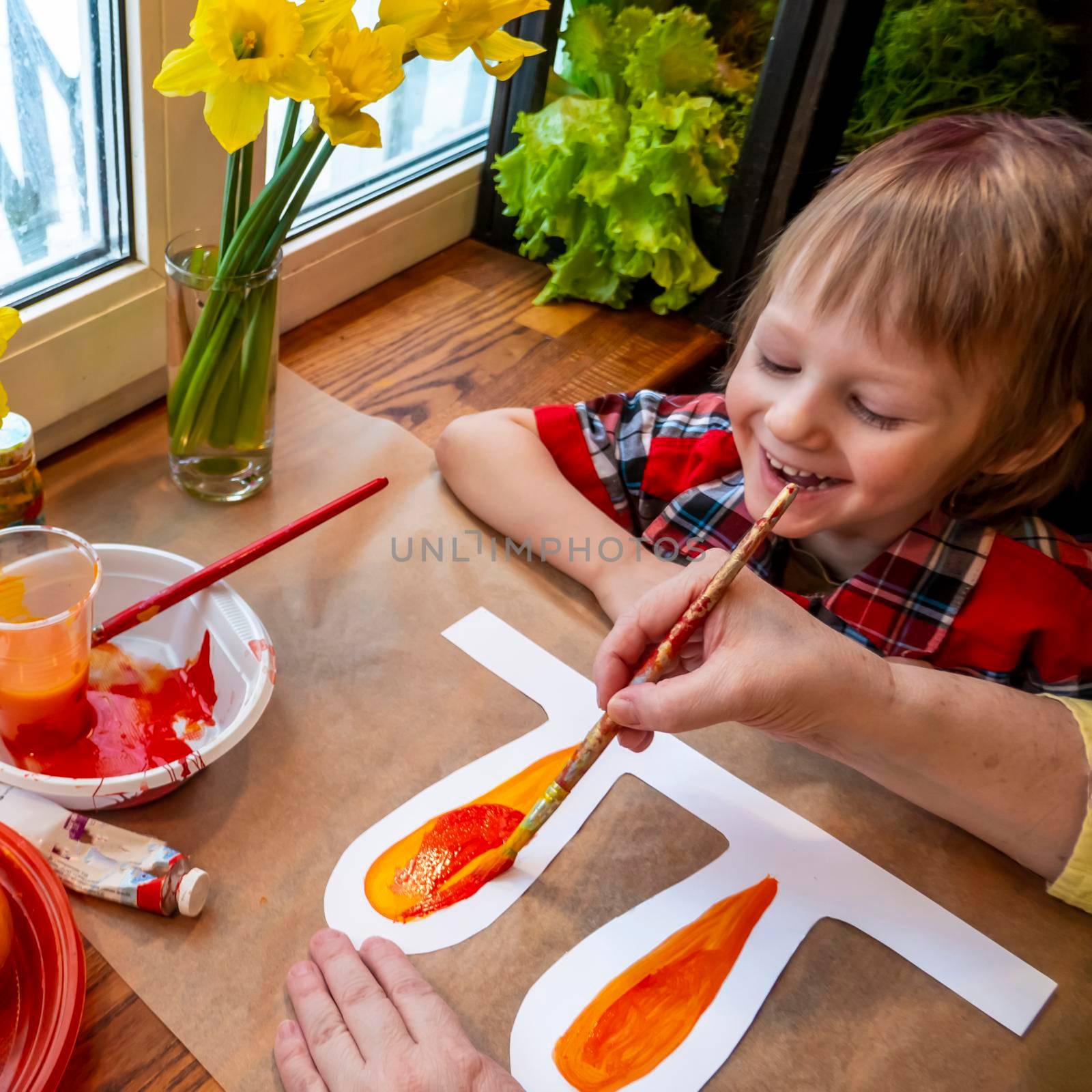 women's speckled hands teach boy draws paper rabbit ears. Preparation for Easter: a child paints paper rabbit ears with paints in the kitchen among fresh greenery, flowers and painted eggs