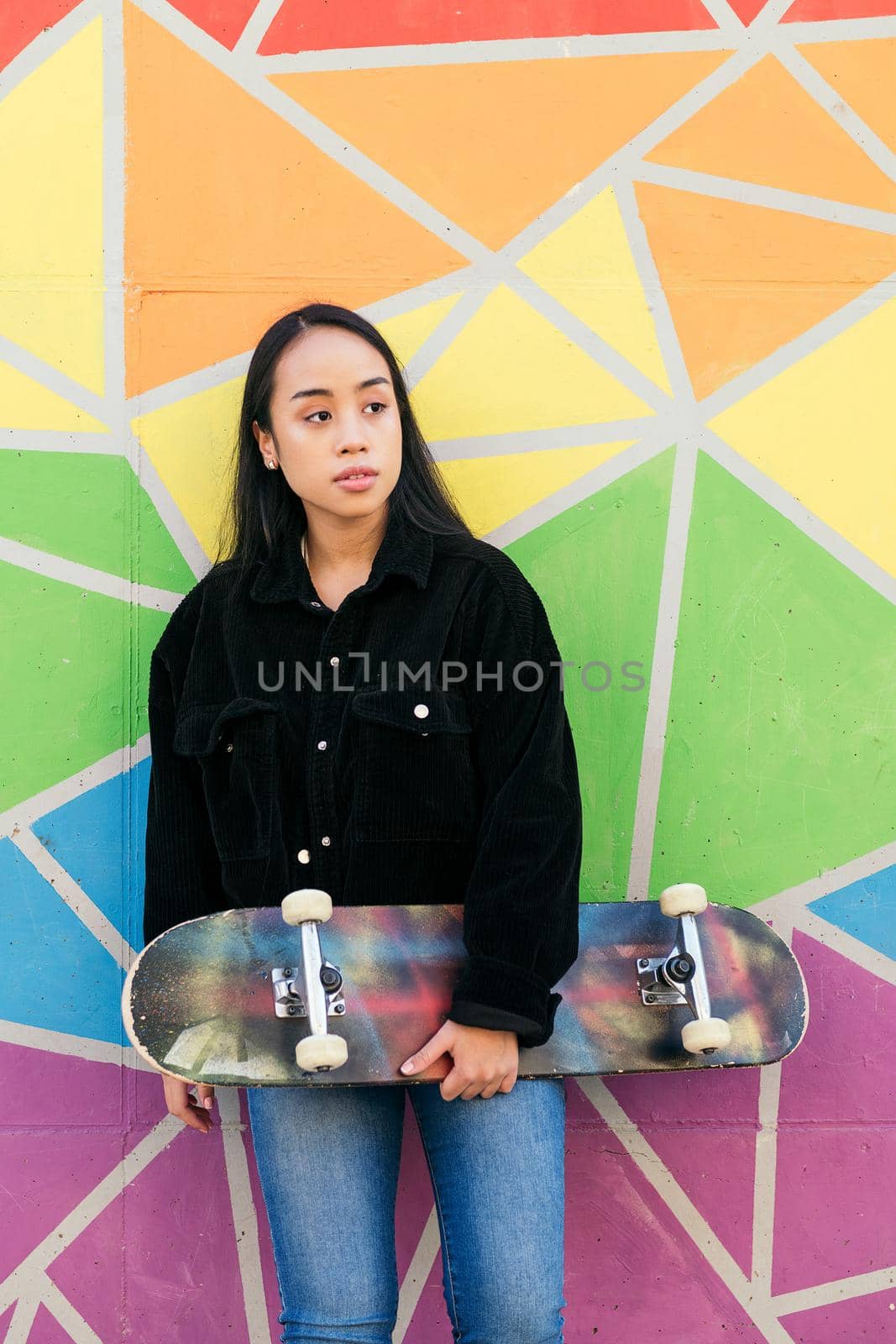young asian woman with her skateboard leaning against a colorful wall