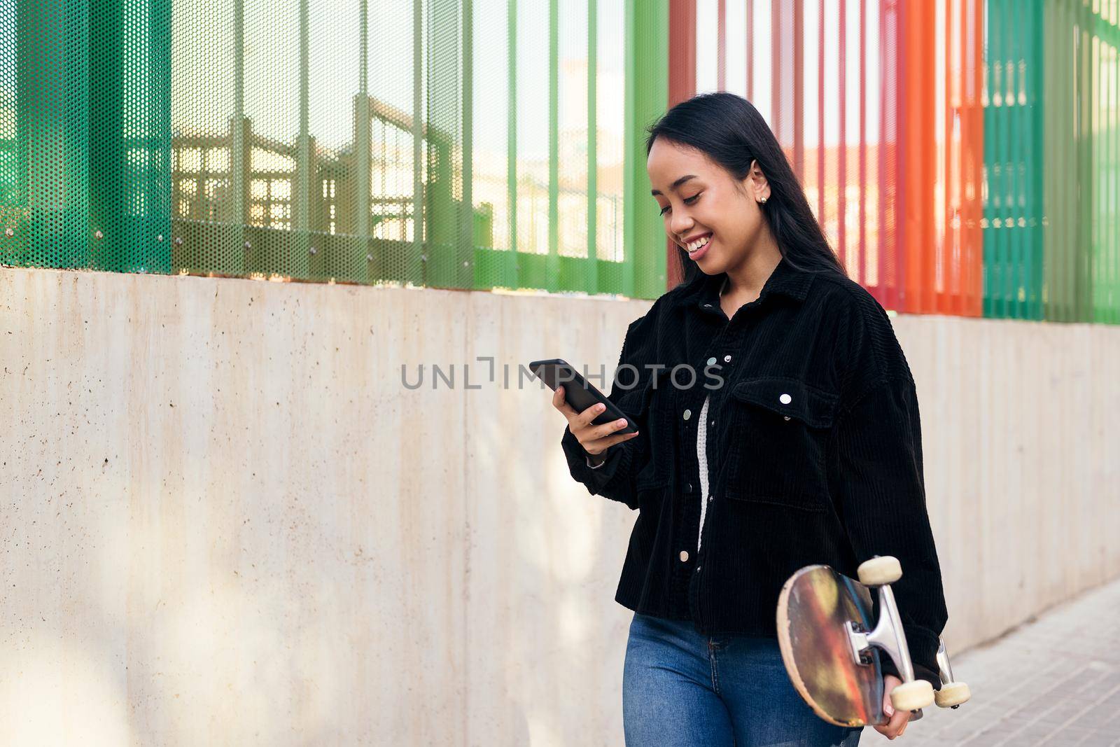 smiling young asian girl typing on the mobile phone while walking down the street with her skateboard in her hand