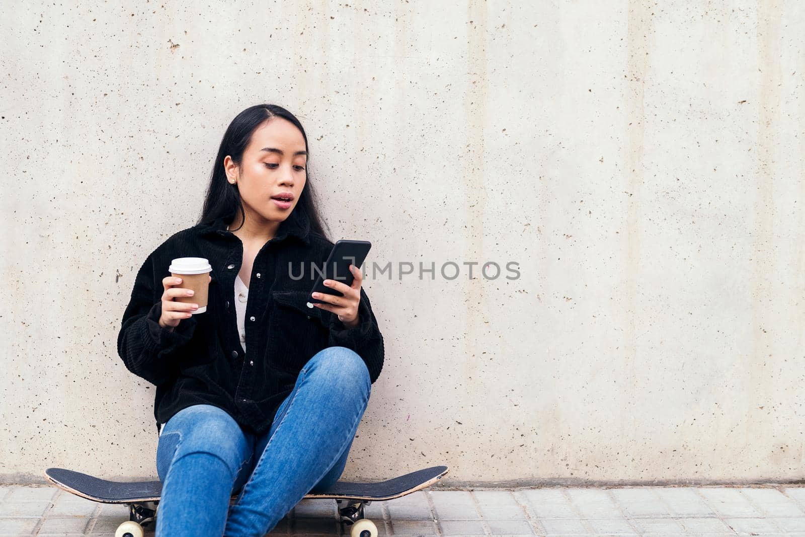 young asian woman typing on a phone sitting outdoors on her skateboard leaning against a concrete wall