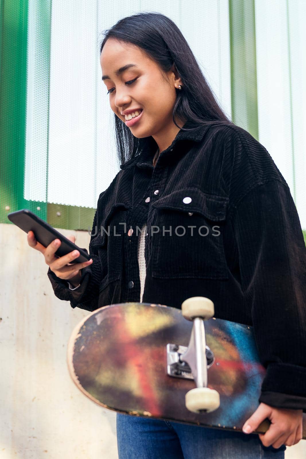 vertical portrait of a smiling young asian woman typing on the mobile phone standing outdoors with her skateboard in her hand