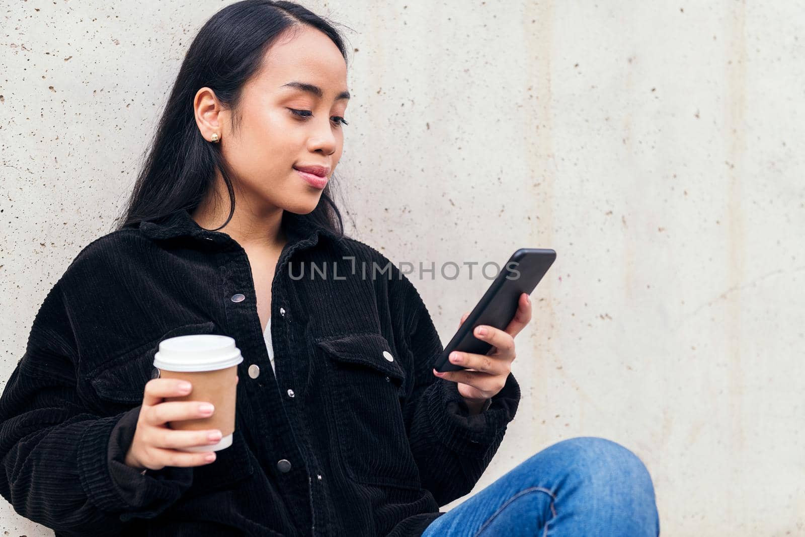 young asian woman with a coffee typing on a phone sitting outdoors leaning against a concrete wall