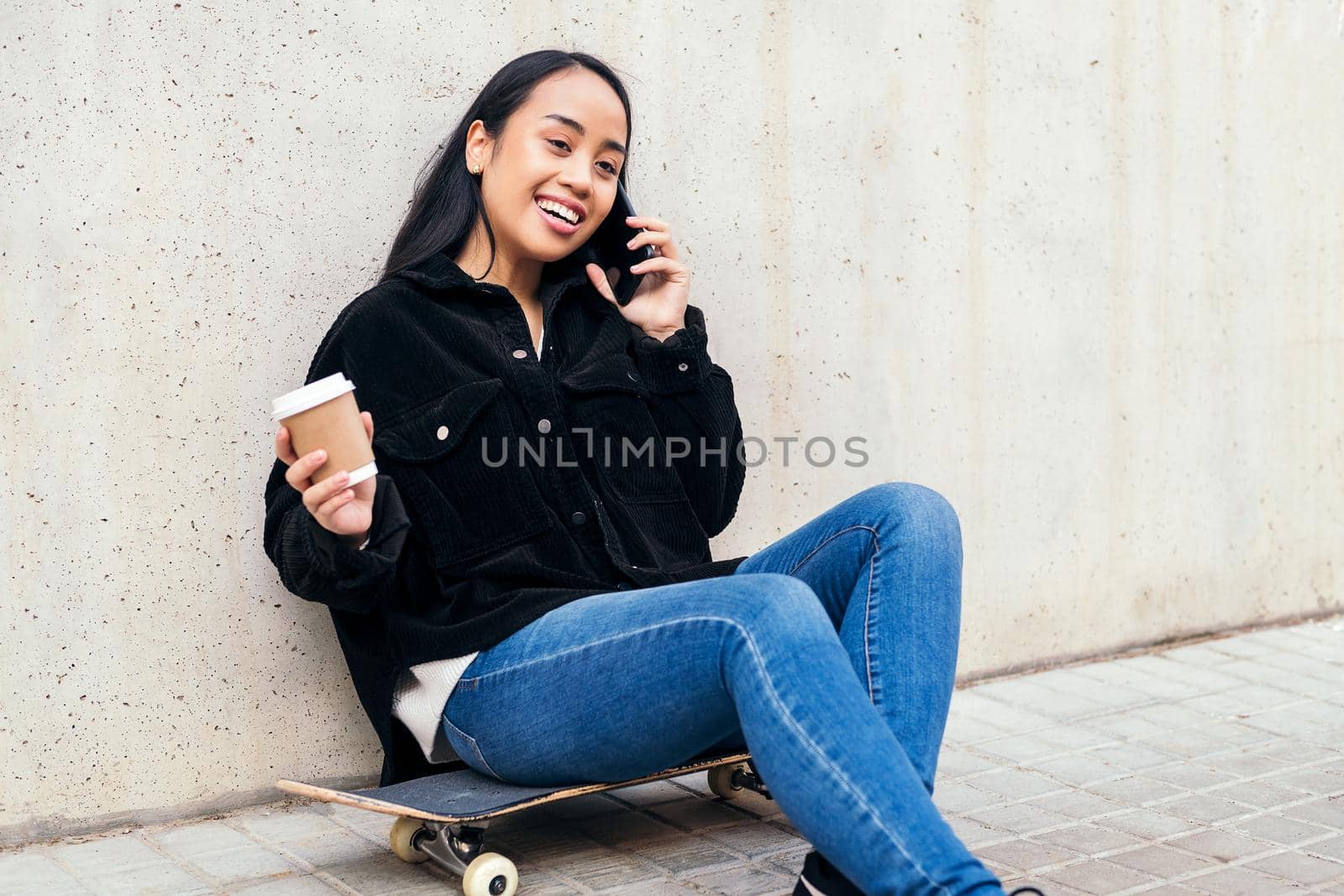 smiling asian girl talking by phone outdoors sitting on her skateboard leaning against a concrete wall