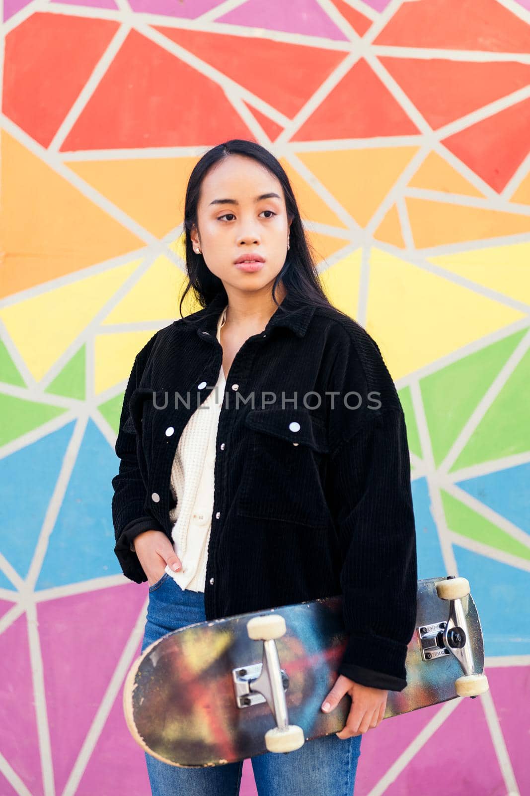 young asian woman with a skateboard in her hand standing in front of a colorful wall