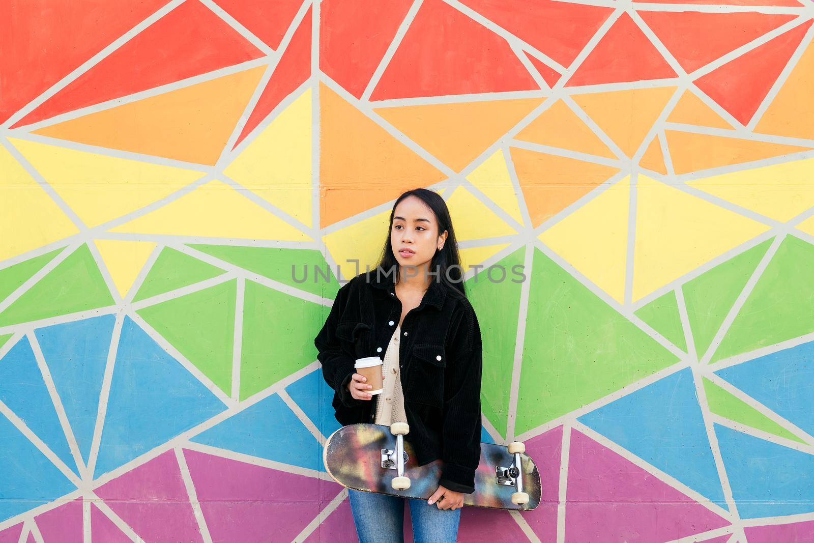 young asian woman with a coffee and a skateboard in her hand leaning in a colorful wall