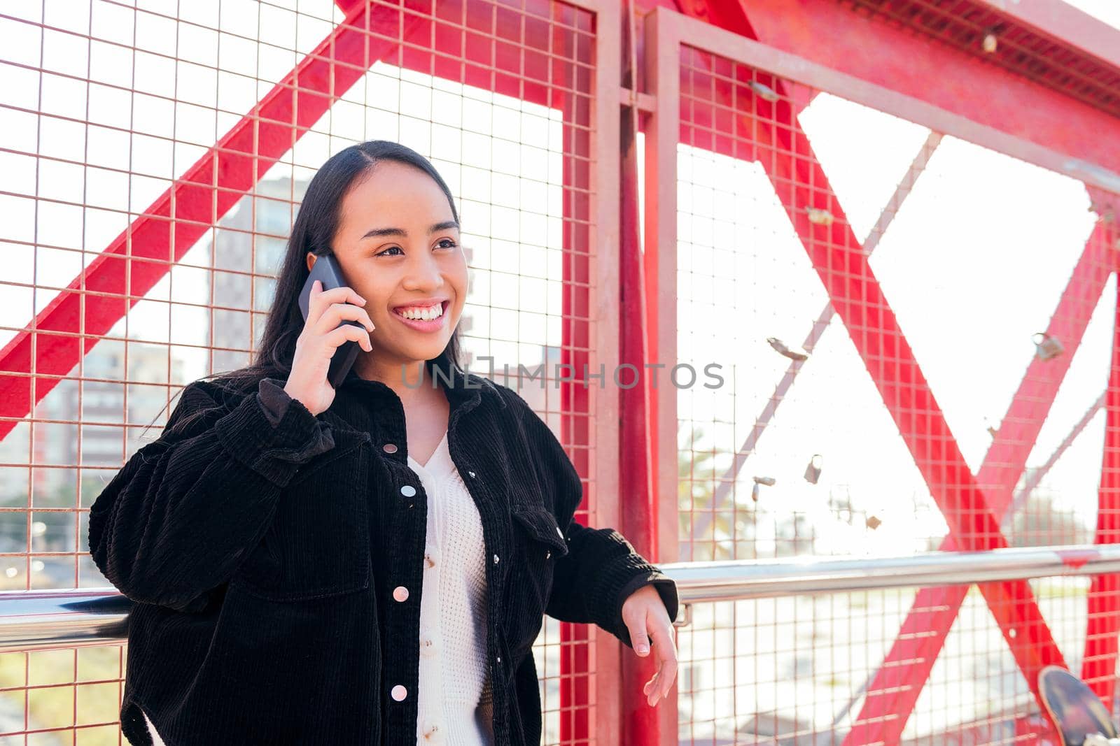 smiling young asian woman outdoors talking by phone leaning against a red fence