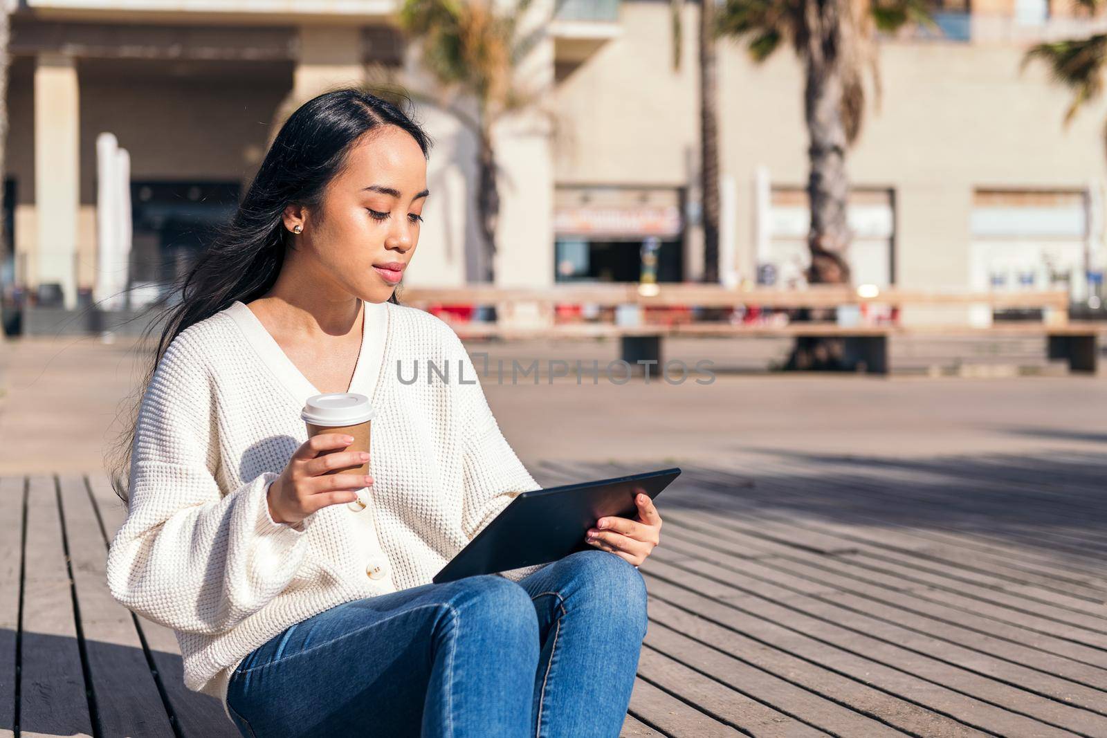 young asian woman relaxing sitting on the street looking at her tablet with a take-away coffee in her hand