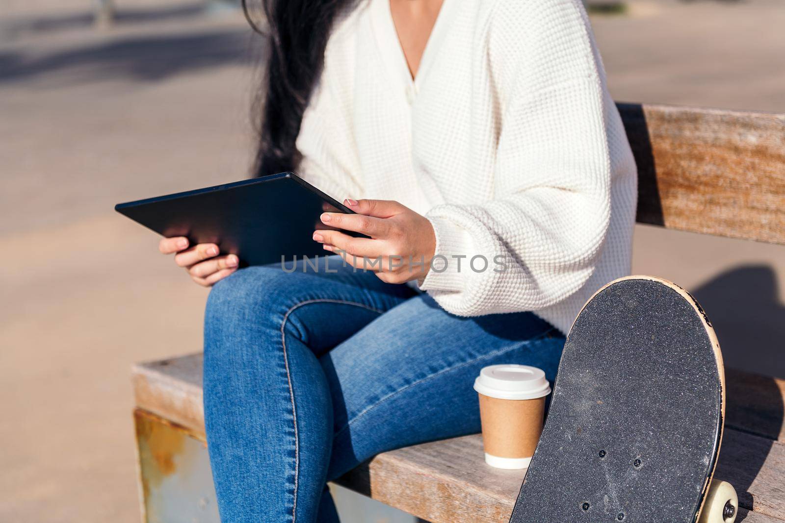 unrecognizable young woman sitting on a bench using her tablet next to a coffee and a skateboard