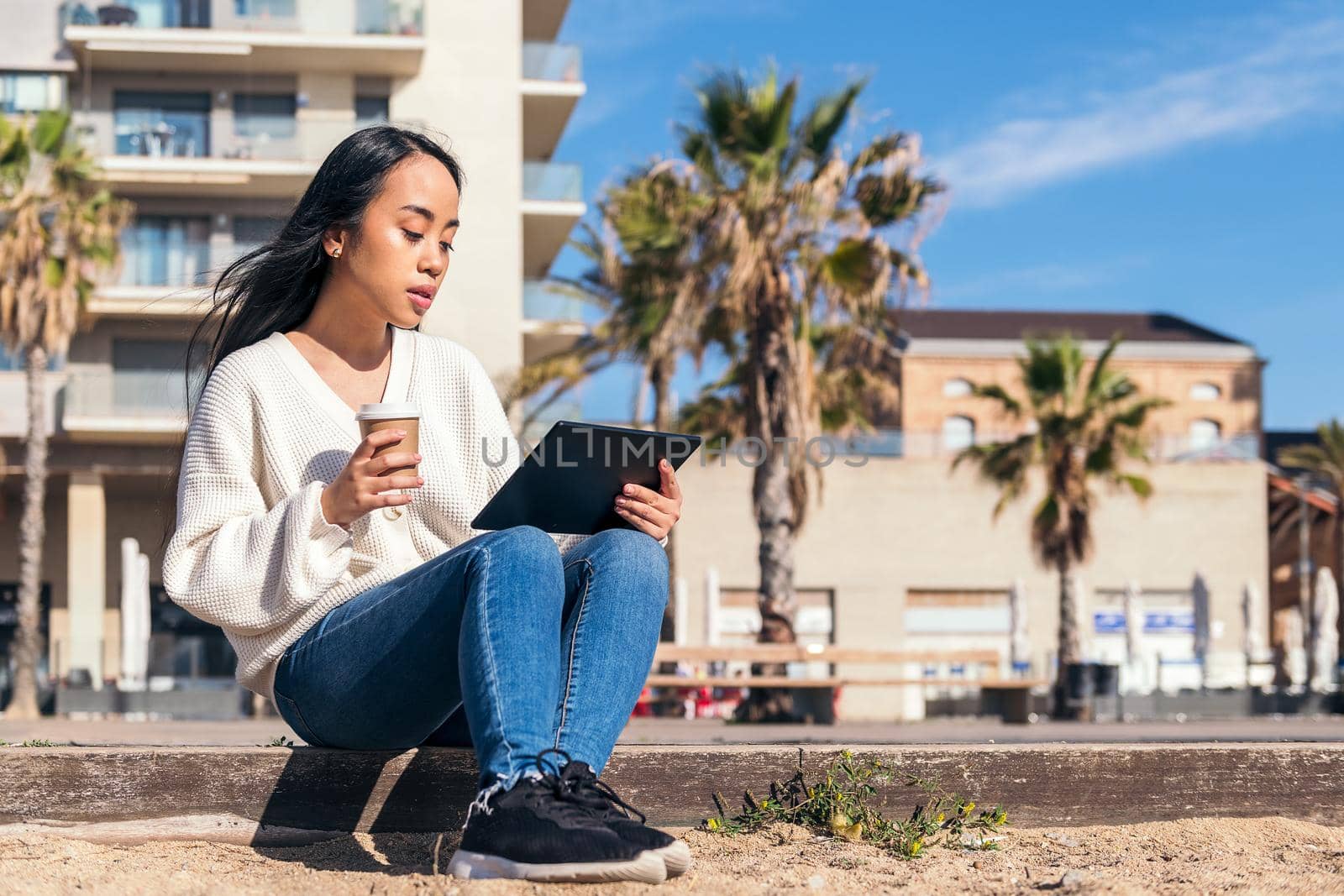 young asian woman relaxing sitting on the street watching her tablet with a take-away coffee in her hand