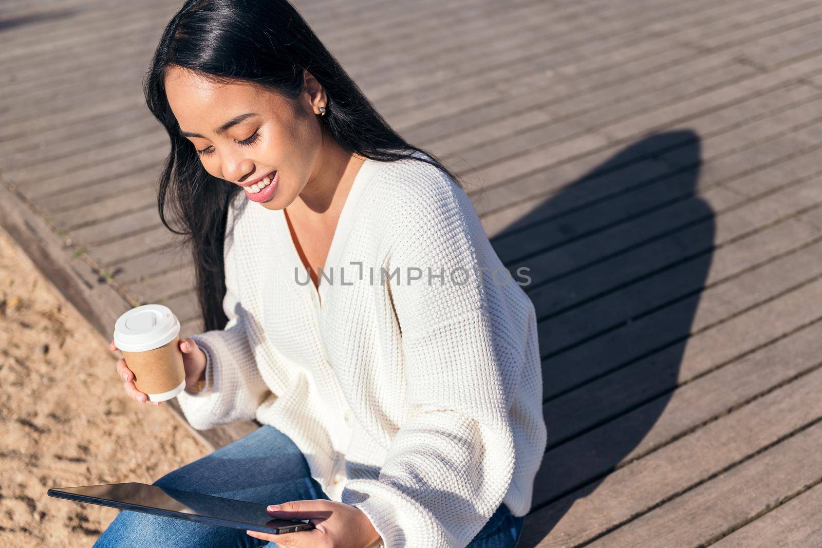 young asian woman smiling sitting outdoors looking at her tablet with a take-away coffee in her hand