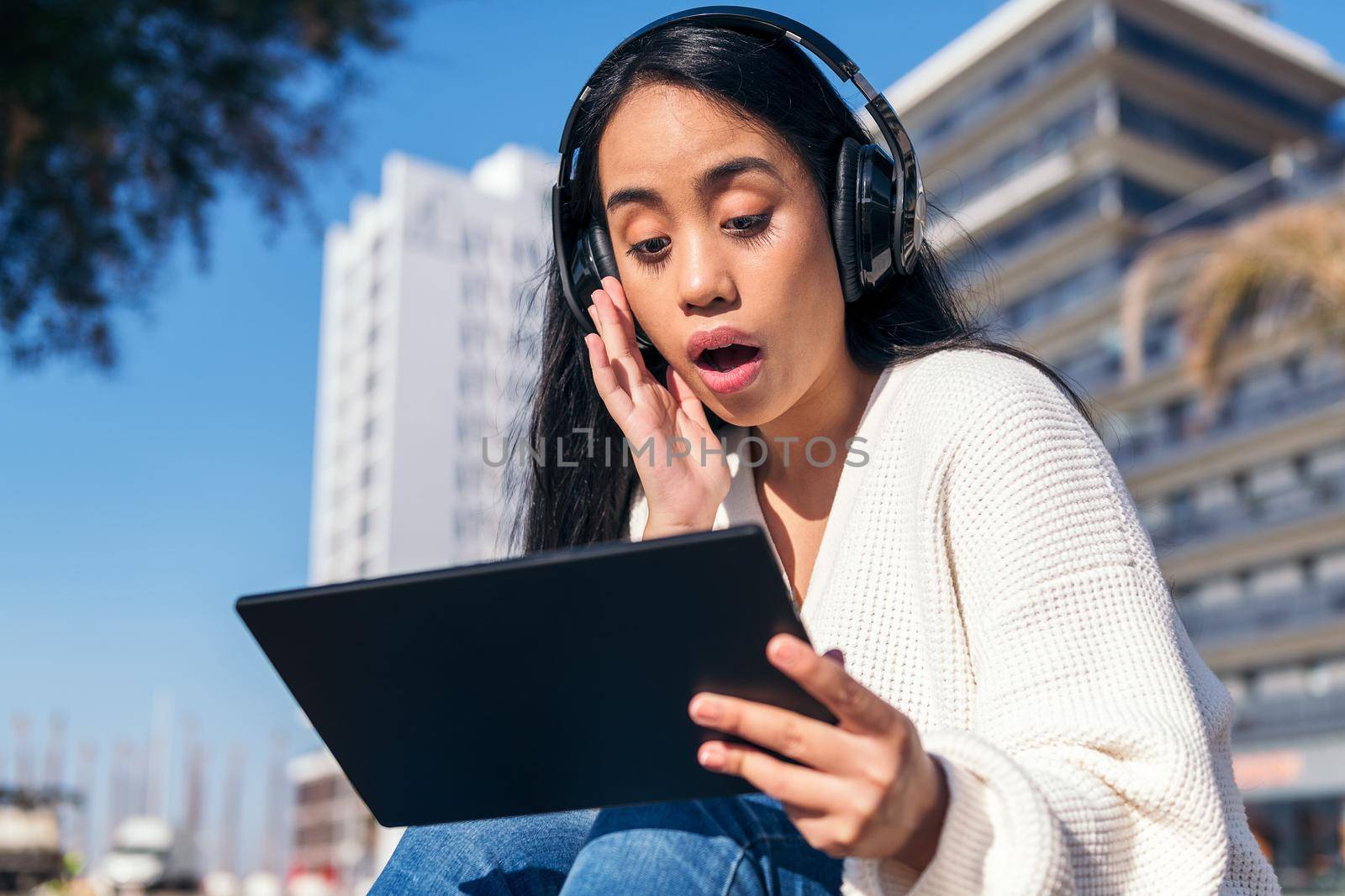 surprised asian young woman with headphones watching videos on her tablet sitting outdoors