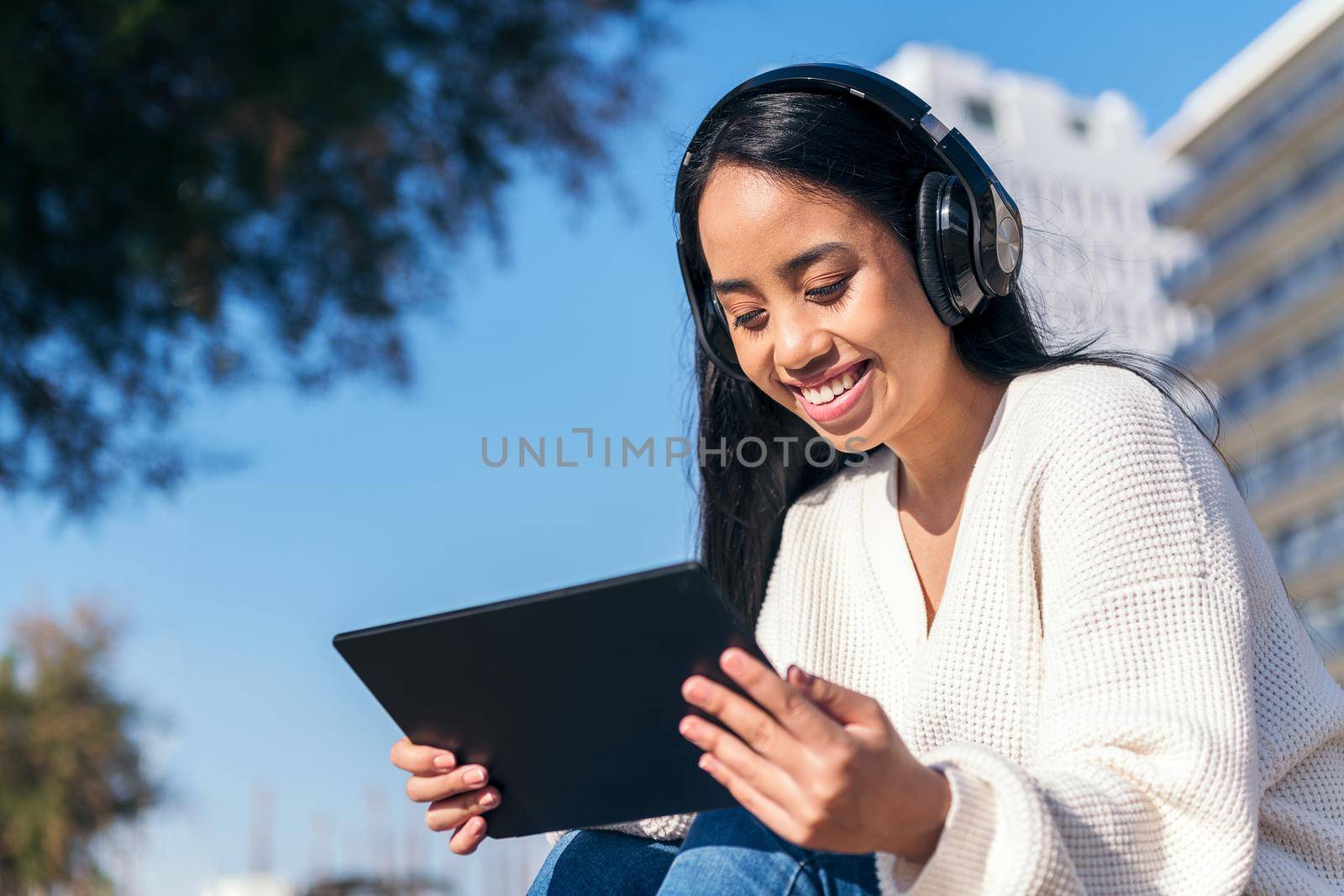 smiling asian young woman with headphones watching videos on her tablet sitting outdoors