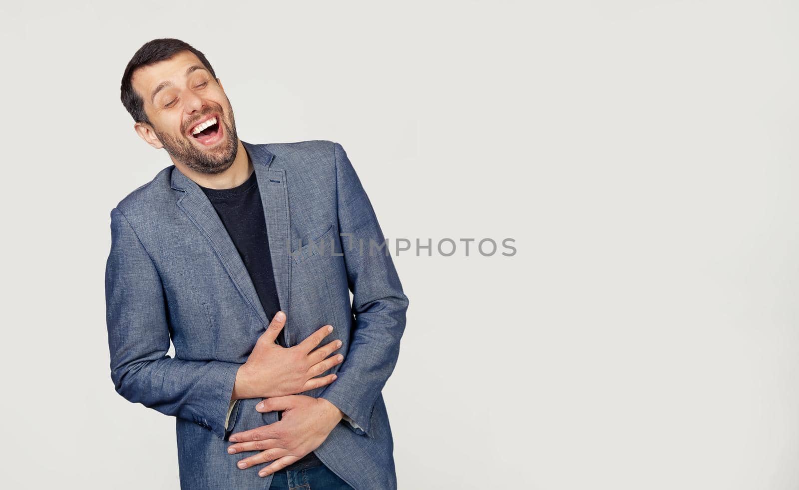 Young businessman man with a beard in a jacket smiling and laughing out loud because a funny crazy joke with hands on the body. Portrait of a man on a gray background.