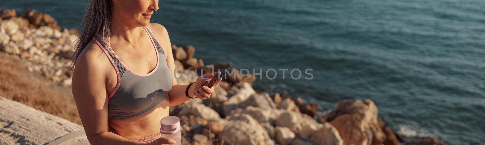 Slender adult woman in sports clothes sitting by the seashore, holding buttle of water and mobile phone