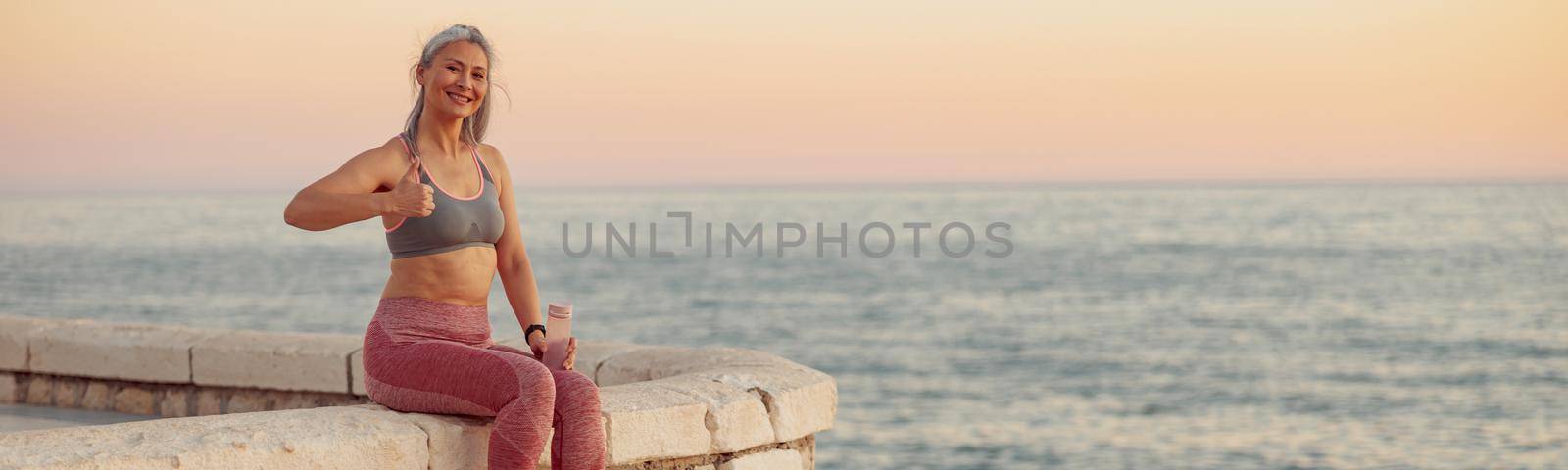 Sporty woman sitting on ledge of waterfront on background with horizon of sea with pink sky, smiling, showing thumbs up