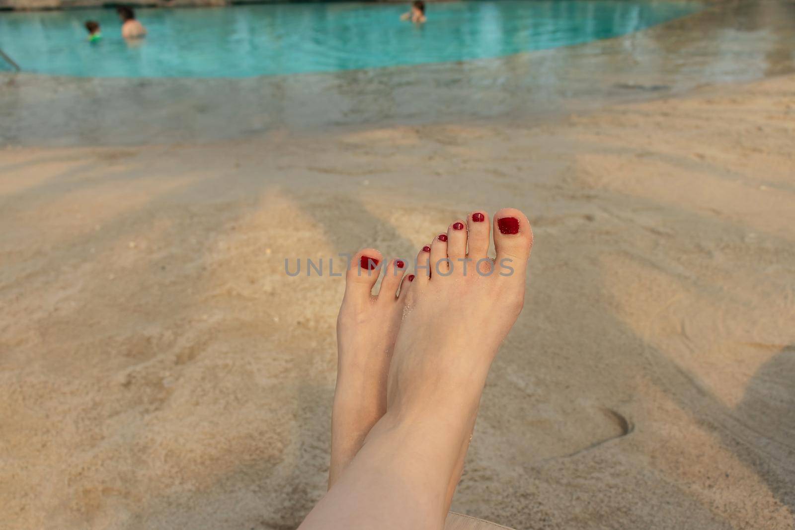 beautiful female feet with red pedicure on the background of blue water and sand on the beach. the concept of relaxation by the sea on a bright, hot and sunny day by Leoschka