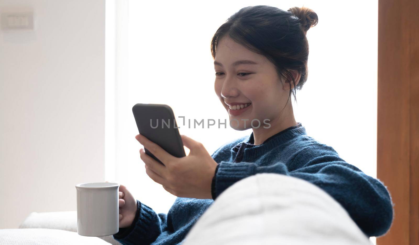 Portrait of a smiling asian women sitting at sofa and drink coffee looking at smartphone.