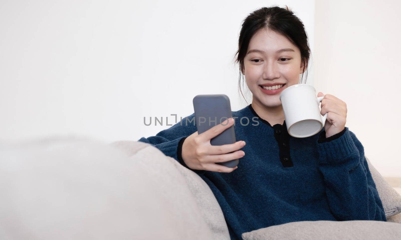 Close-up Of Smiling thailand Lady Having Cellphone Conversation And Enjoying Hot Drink While Relaxing On Couch. by wichayada
