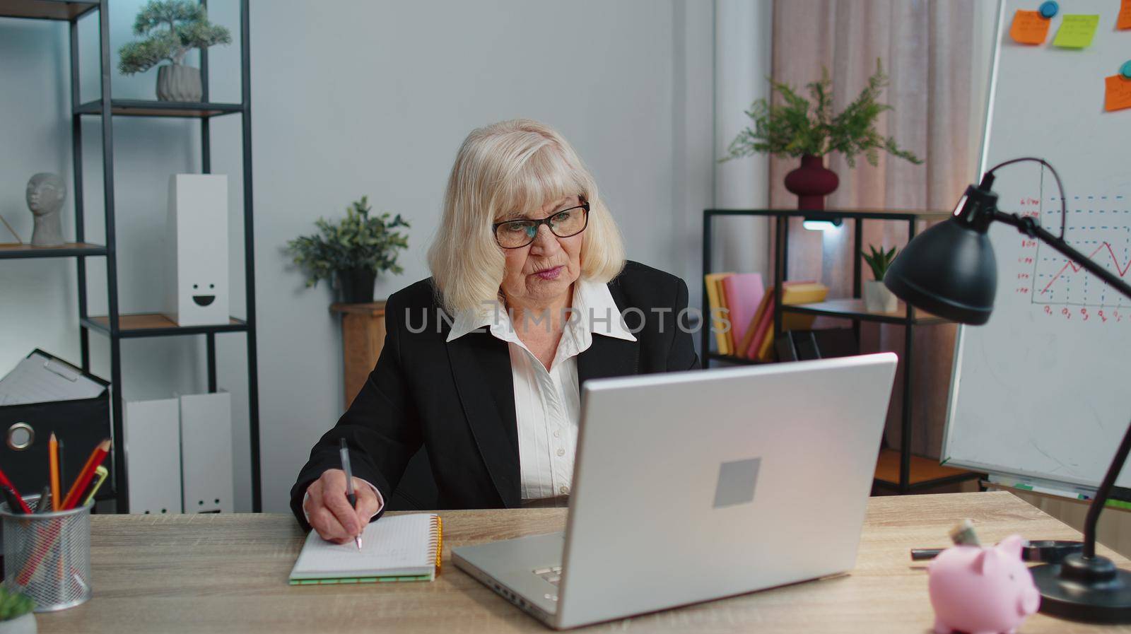 Senior mature older business woman elearning online concept, watching business training class, live webinar on laptop computer remote working, social distance learning, writing notes from home office