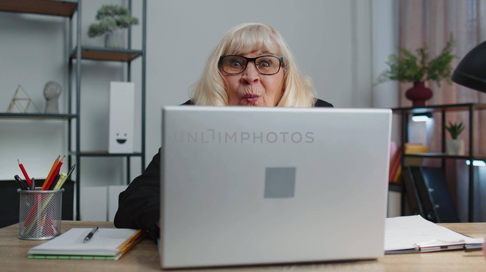 Senior mature older business woman hiding behind laptop computer, making funny face, fooling around, putting her tongue out remote working disrespecting someone at home office. Elderly grandmother