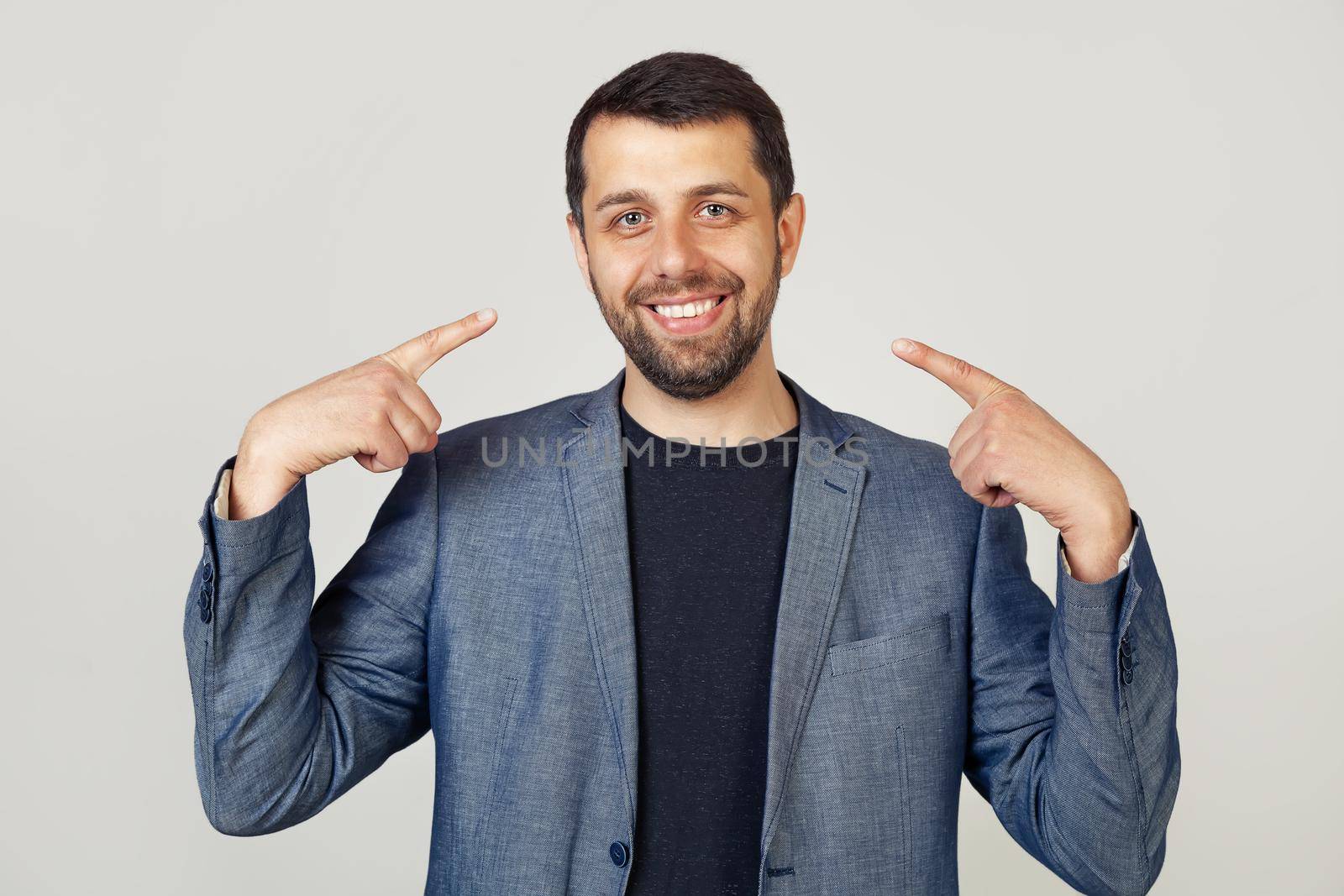 Young businessman man with beard in jacket smiling confidently showing and pointing with fingers teeth and mouth. Health concept. Portrait of a man on a gray background.