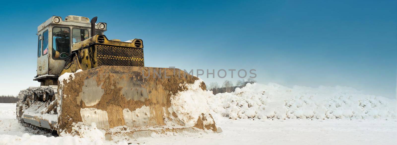 Yellow crawler tractor bulldozer with a bucket works in winter clearing the road from snow. Frosty day with bright sun and beautiful blue sky in the background. Yellow bulldozer in winter