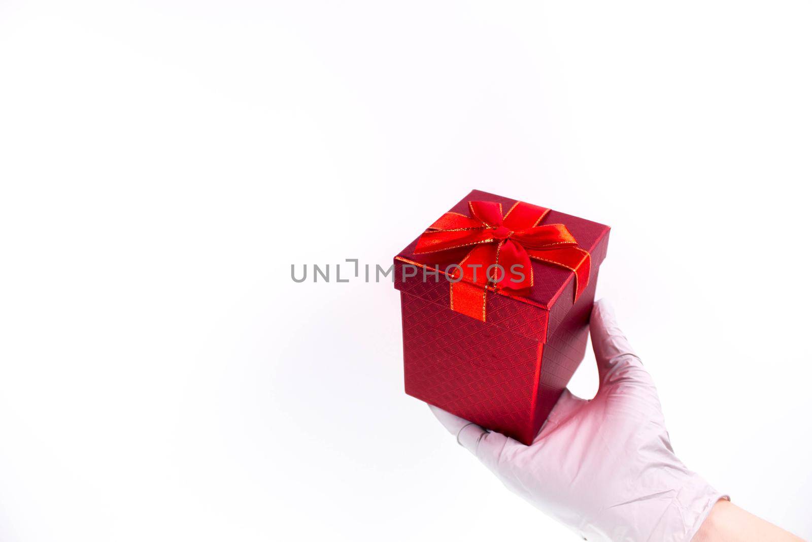 Women's hand in medical gloves hold a gift box with a red ribbon on a white background. Concept of sales and gifts during Coronavirus. Quarantine. Delivery courier. Online purchase