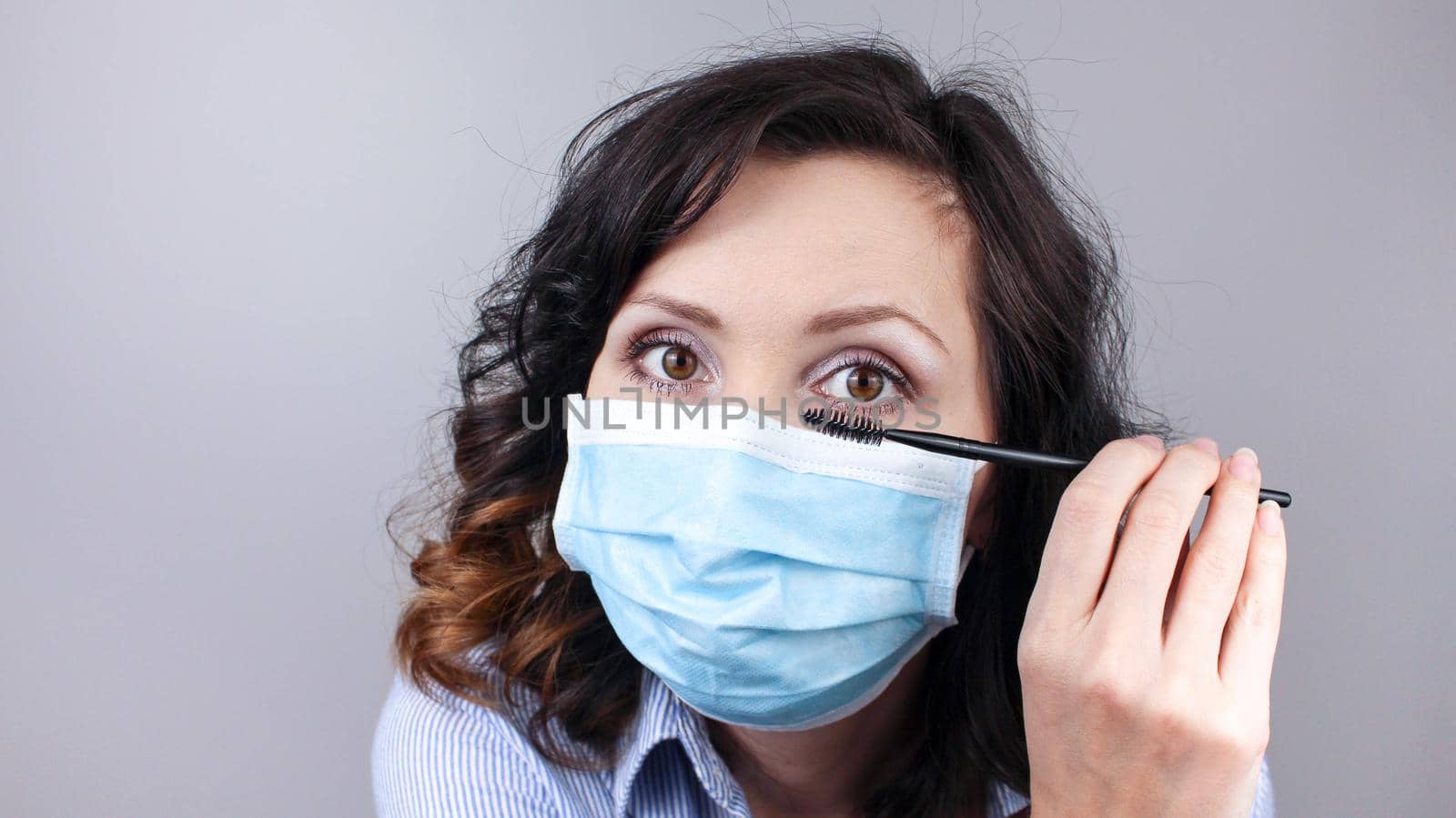 Woman wearing protection face mask against coronavirus and applying make-up. Woman in a mask looking in a mirror and applying mascara. Medical mask, Close up shot, Prevention from covid19