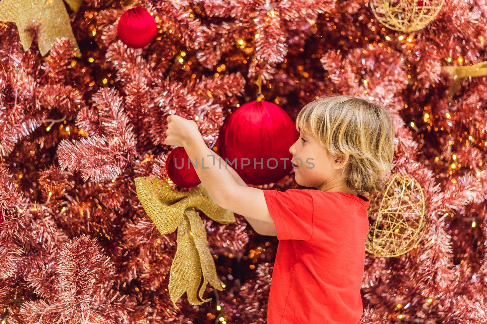 Boy hangs a decoration on the red Christmas tree. Red Christmas tree on the background of lights.