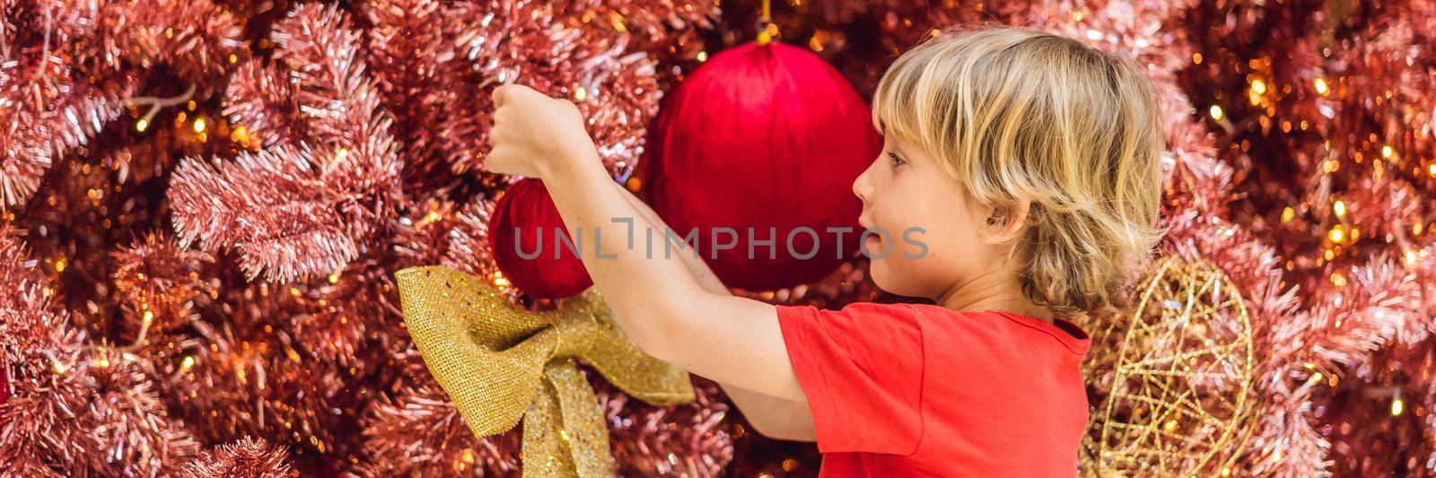 Boy hangs a decoration on the red Christmas tree. Red Christmas tree on the background of lights. BANNER, LONG FORMAT