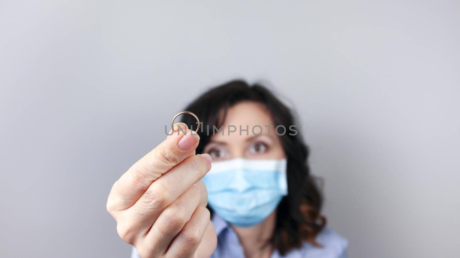 Woman in protective mask holding wedding ring against light background, focus on hand. Divorce during coronavirus quarantine