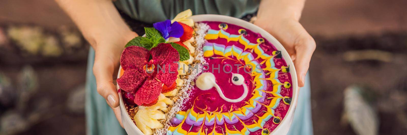 BANNER, LONG FORMAT Young woman having a mediterranean breakfast, eats Healthy tropical breakfast, smoothie bowl with tropical fruits, decorated with a pattern of colorful yogurt with turmeric and spirulina. It is also decorated with fruits, flowers, chia seeds, coconut, granola, pineapple, mint and strawberries by galitskaya