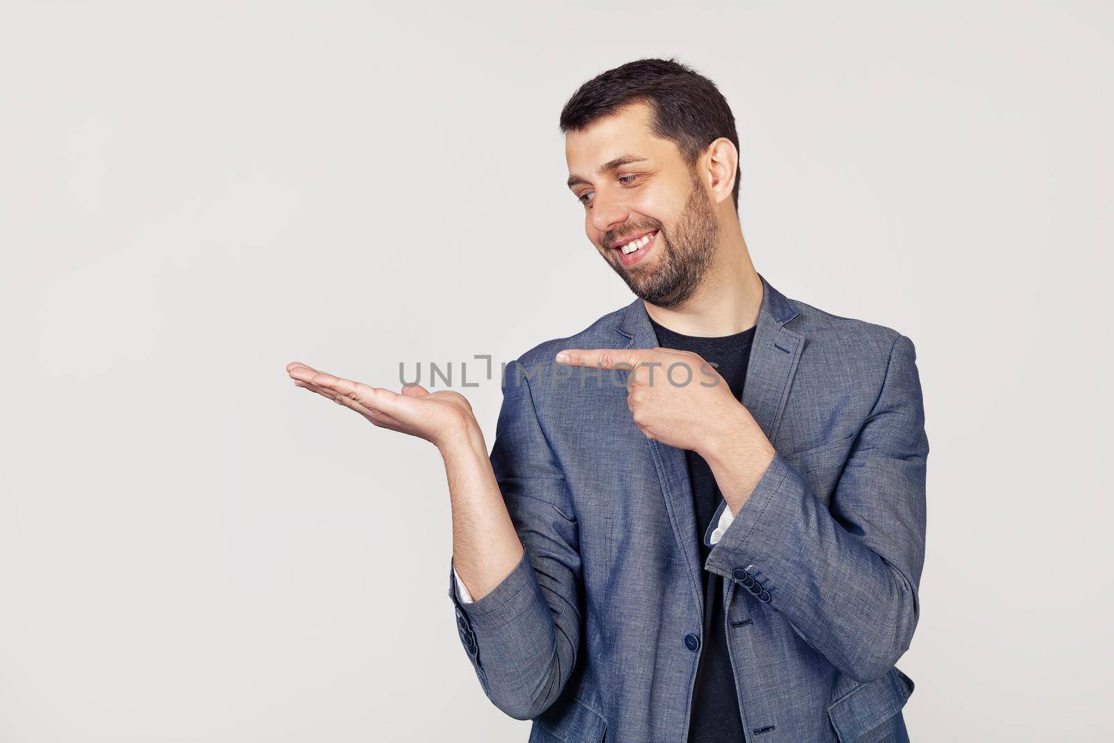 Young businessman man with a beard in a jacket looks at the palm, showing with his hand and pointing his finger. Portrait of a man on a gray background by ViShark