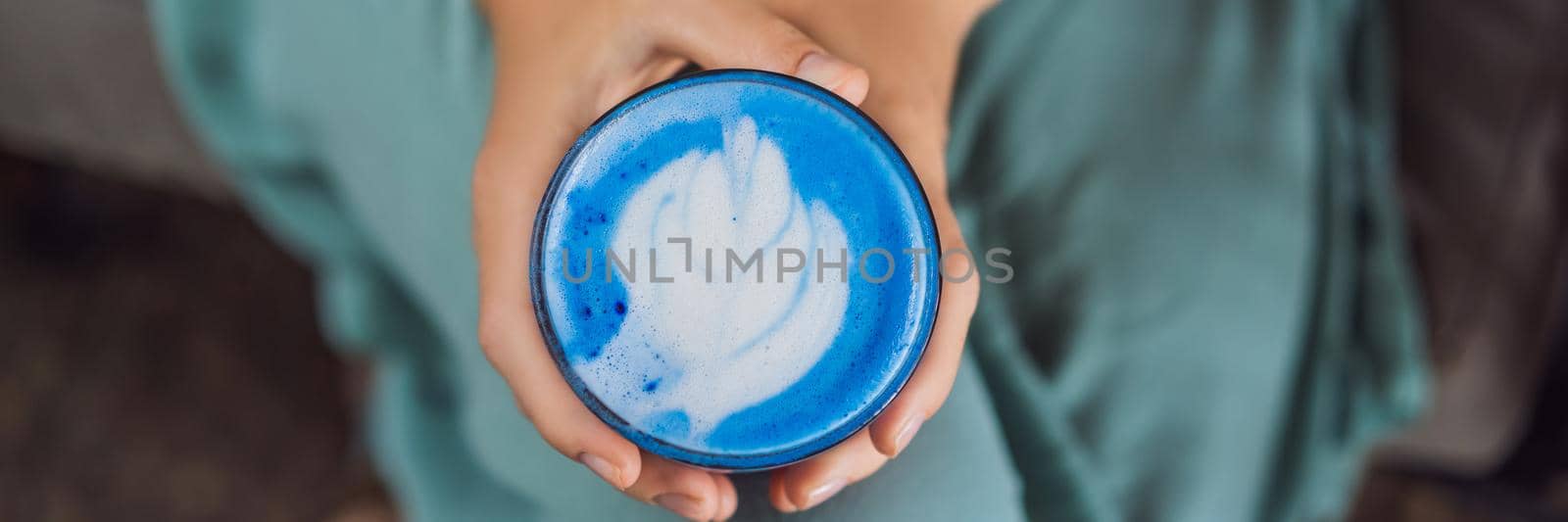 Young woman having a mediterranean breakfast seated at sofa and drinks Trendy drink: Blue latte. Hot butterfly pea latte or blue spirulina latte BANNER, LONG FORMAT by galitskaya